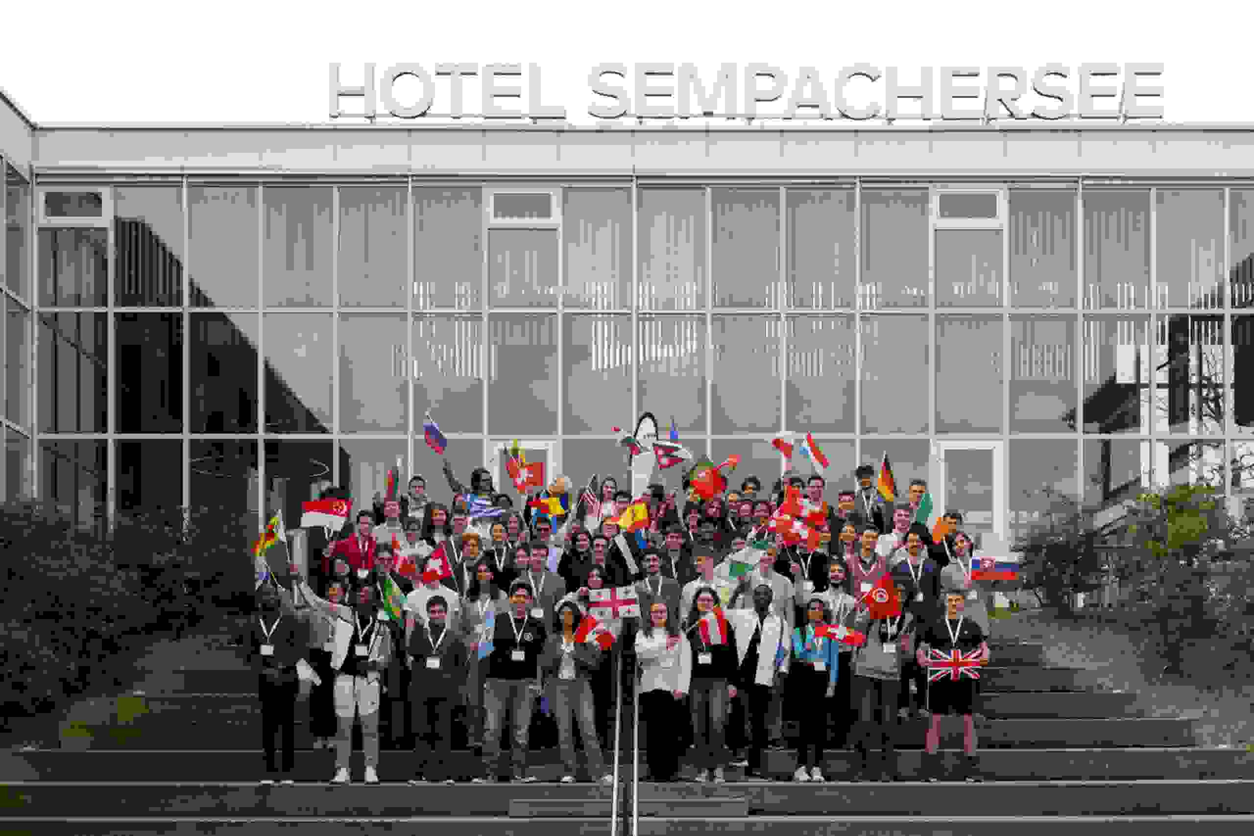 Internationale Teilnehmende des Swiss Talent Forums 2024 vor dem Hotel Sempachersee mit Flaggen aus aller Welt. Gruppenfoto, Hotel Sempachersee, Flaggen, globale Teilnehmende, markenerlebnis, Brand Experience, live kommunikation, schweizer Jugend forscht
