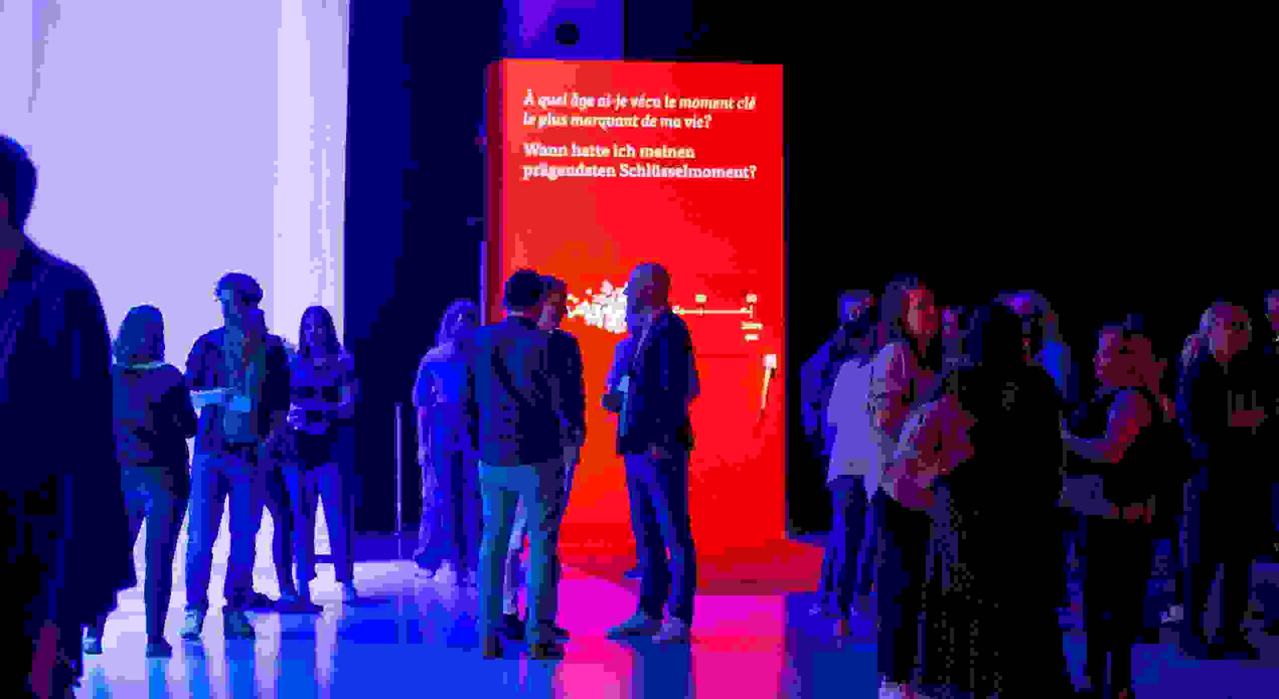 Besucher interagieren bei einem  vor einer auffälligen roten Erlebnisinstallation mit der Frage nach prägenden Schlüsselmomenten. Eventtechnik, live kommunikation, markenerlebnis, Brand Experience, interaktive Ausstellung, Eventbeleuchtung, Corporate-Event