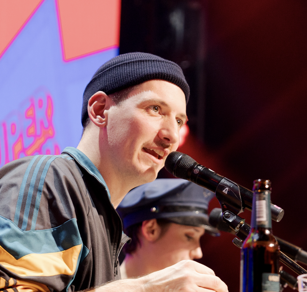 Moderator Mathias Schenk mit Mikrofon und Mützet führt durch das Programm – humorvolle Bühnenmoderation mit Überraschungselementen. Bühnenmoderation, Publikumsnähe, Eventhumor, Supermercado, Show, eventagentur bern
eventplanung, Eventtechnik, live kommunikation, teamevents, Corporate-Event, bernexpo