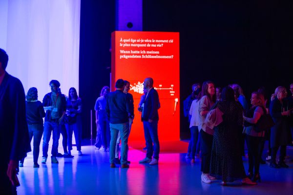 Besucher interagieren bei einem  vor einer auffälligen roten Erlebnisinstallation mit der Frage nach prägenden Schlüsselmomenten. Eventtechnik, live kommunikation, markenerlebnis, Brand Experience, interaktive Ausstellung, Eventbeleuchtung, Corporate-Event