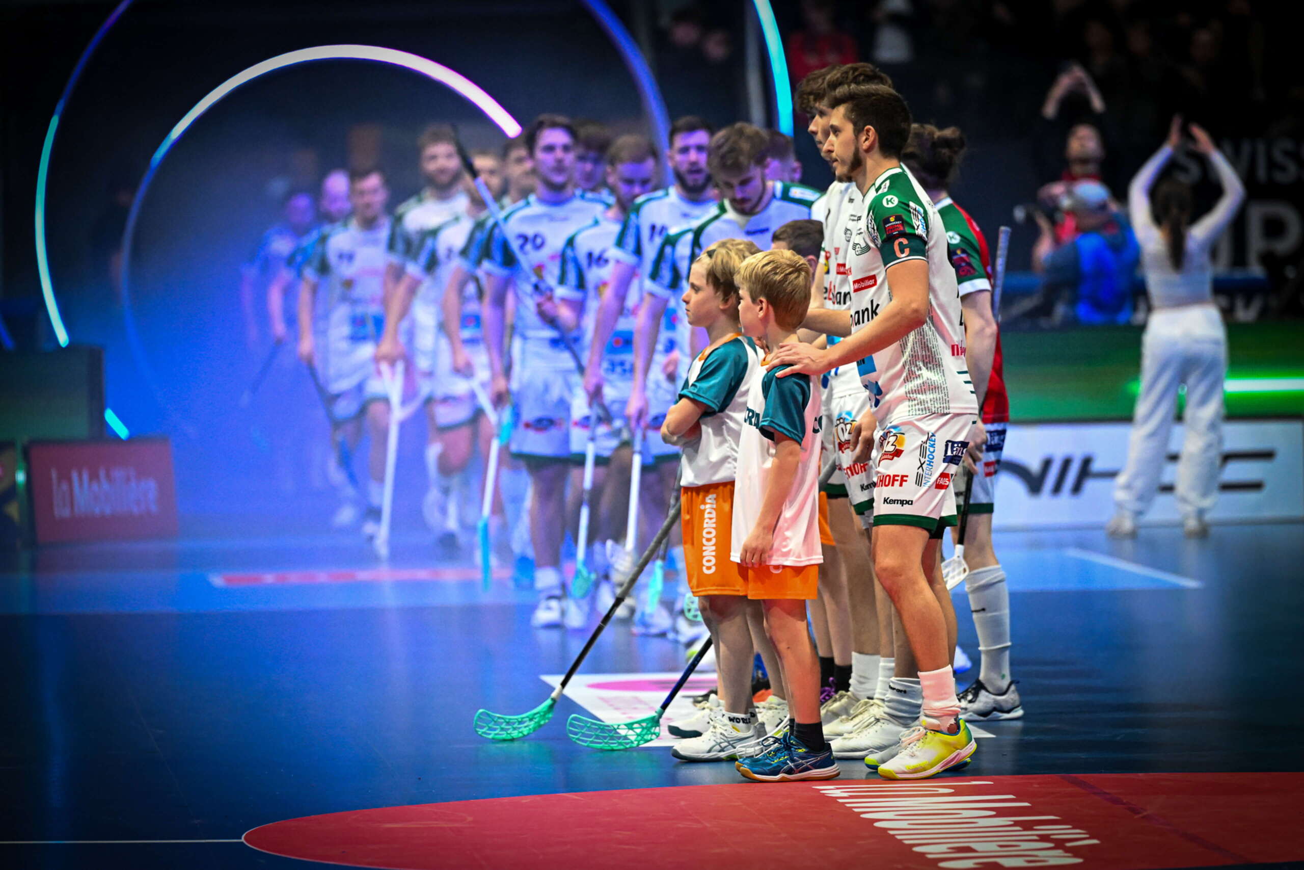 evenjo_superfinal_Swiss_unihockey_sport-event_einlaufen.jpg Das Team bereitet sich mit Einlaufkindern auf den Superfinal vor. konzentrierte Stimmung vor dem Anpfiff unter Lichtinszenierung.Unihockey Superfinal, Teampräsentation, Einlaufkinder, Sportevent, Floorball Schweiz, Lichtshow, Spielbeginn, live kommunikation, Corporate-Event, Eventagentur bern, eventplanung, firmenevents, teamevents bern, jubiläumsfeier, networking anlass, mitarbeiteranlass, evenjo-Team, Technik, Public Events, Sport und Entertainment Events, markenerlebnis, Brand Experience, Corporate-Event, Lichtshow, Eröffnungsshow
