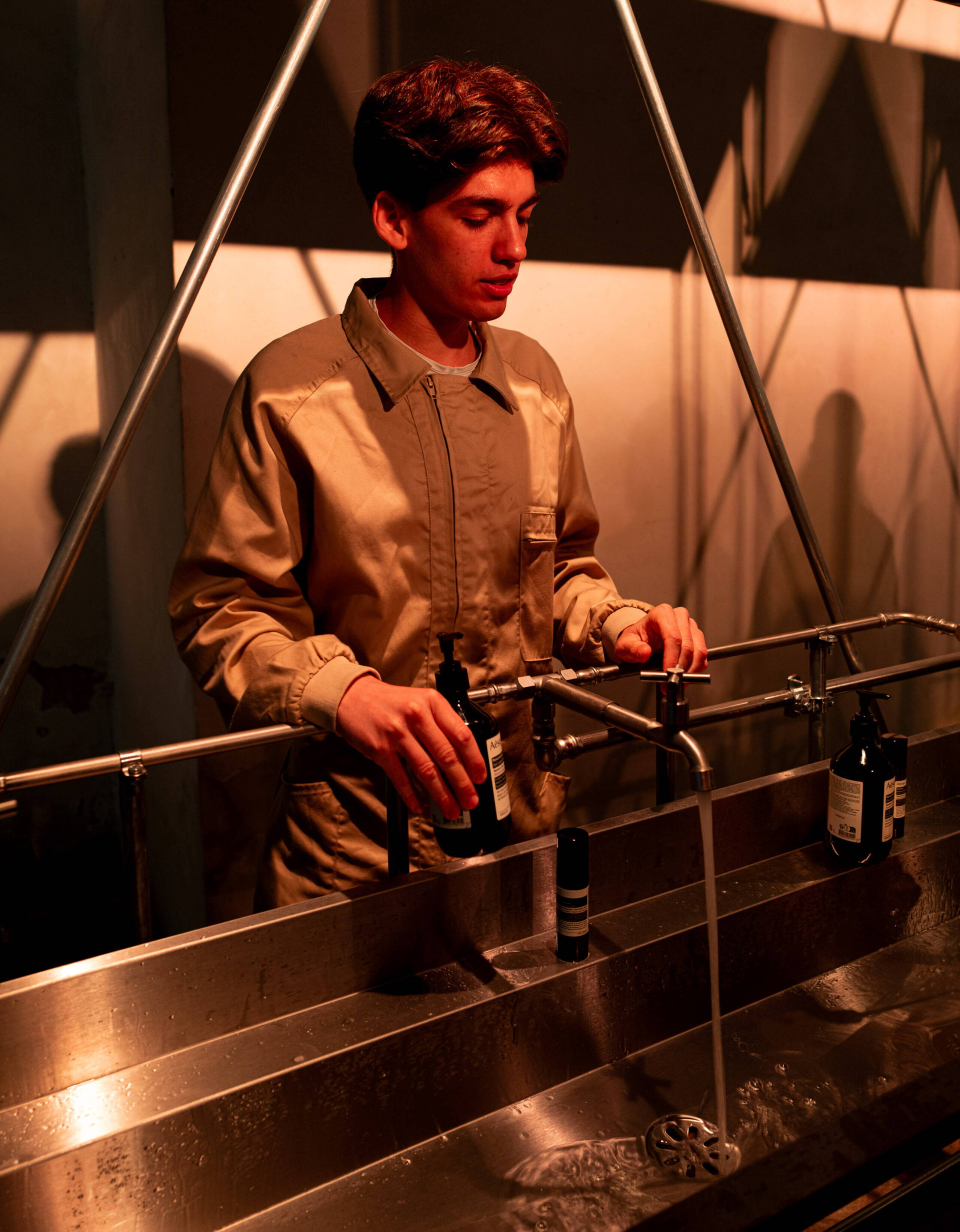 Warmes, rötliches Licht beleuchtet einen Mitarbeitenden der Aesop-Installation an der Milan Design Week in Milano. Er steht an einer langen Edelstahl-Waschstation mit fliessendem Wasser und bereitet Produkte für die Besucher:innen vor.