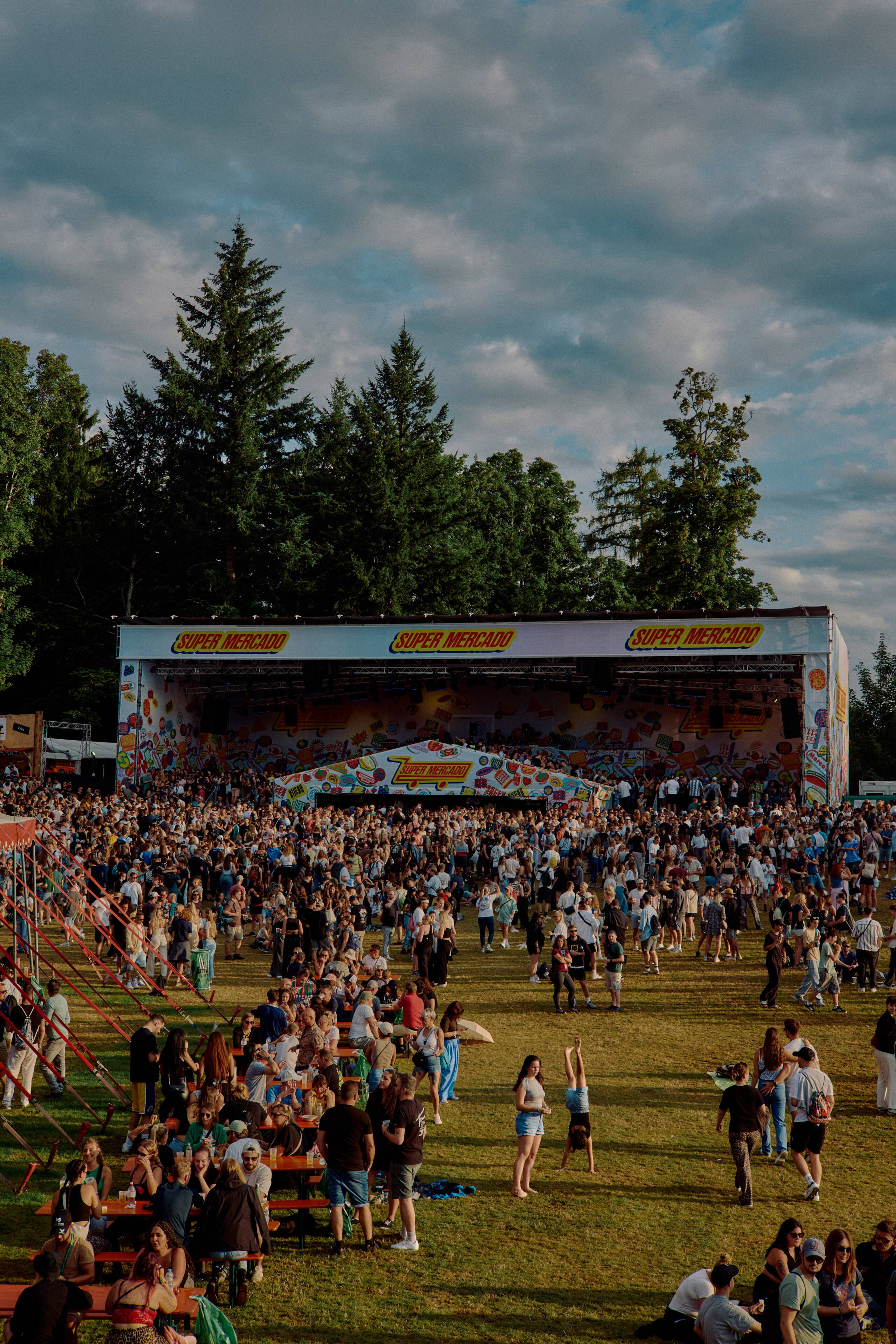 gurtenfestival-evenjo-ag-bern_Supermercado.jpg Festivalbesucher:innen stehen und sitzen tagsüber vor der „Supermercado“-Bühne beim Gurtenfestival in Bern. Im Vordergrund sind Picknicktische, im Hintergrund die Bühne mit bunten Grafiken.