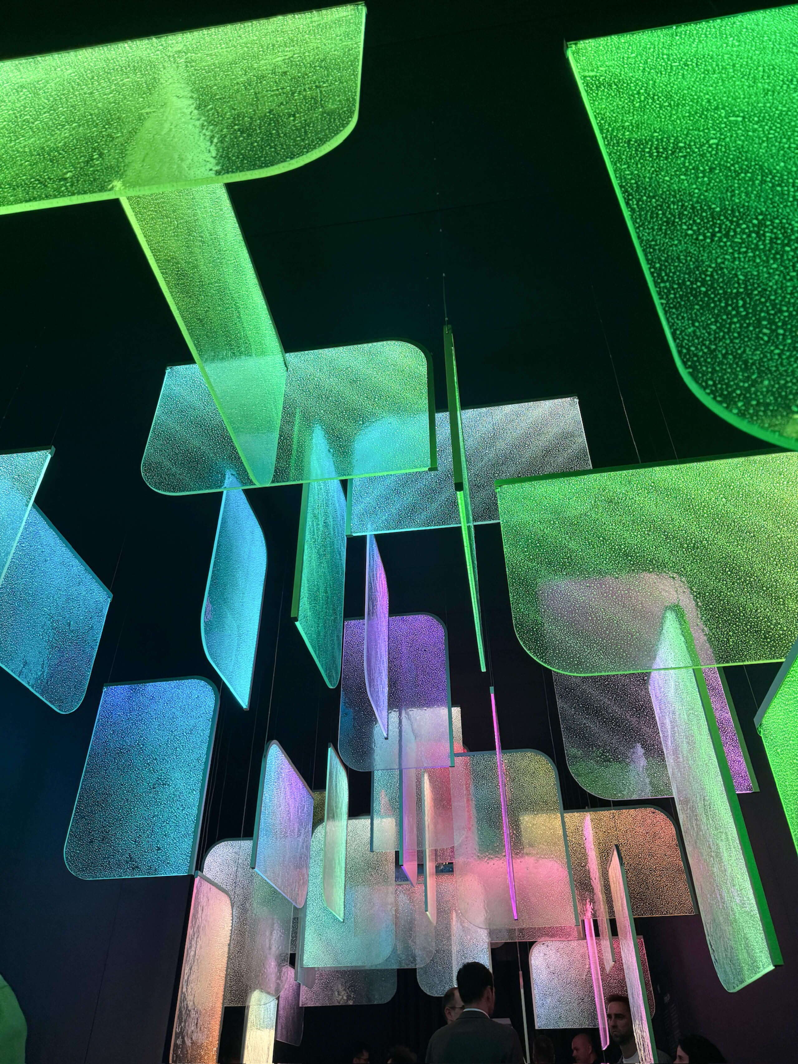 Besucher:innen stehen unter der Lichtinstallation «Drifting Lights» von Preciosa am Fuorisalone der Milan Design Week in Milano. Transparente, leuchtende Elemente in Grün, Blau und Rosa schweben von der Decke und erzeugen eine immersive, fast futuristische Atmosphäre.
