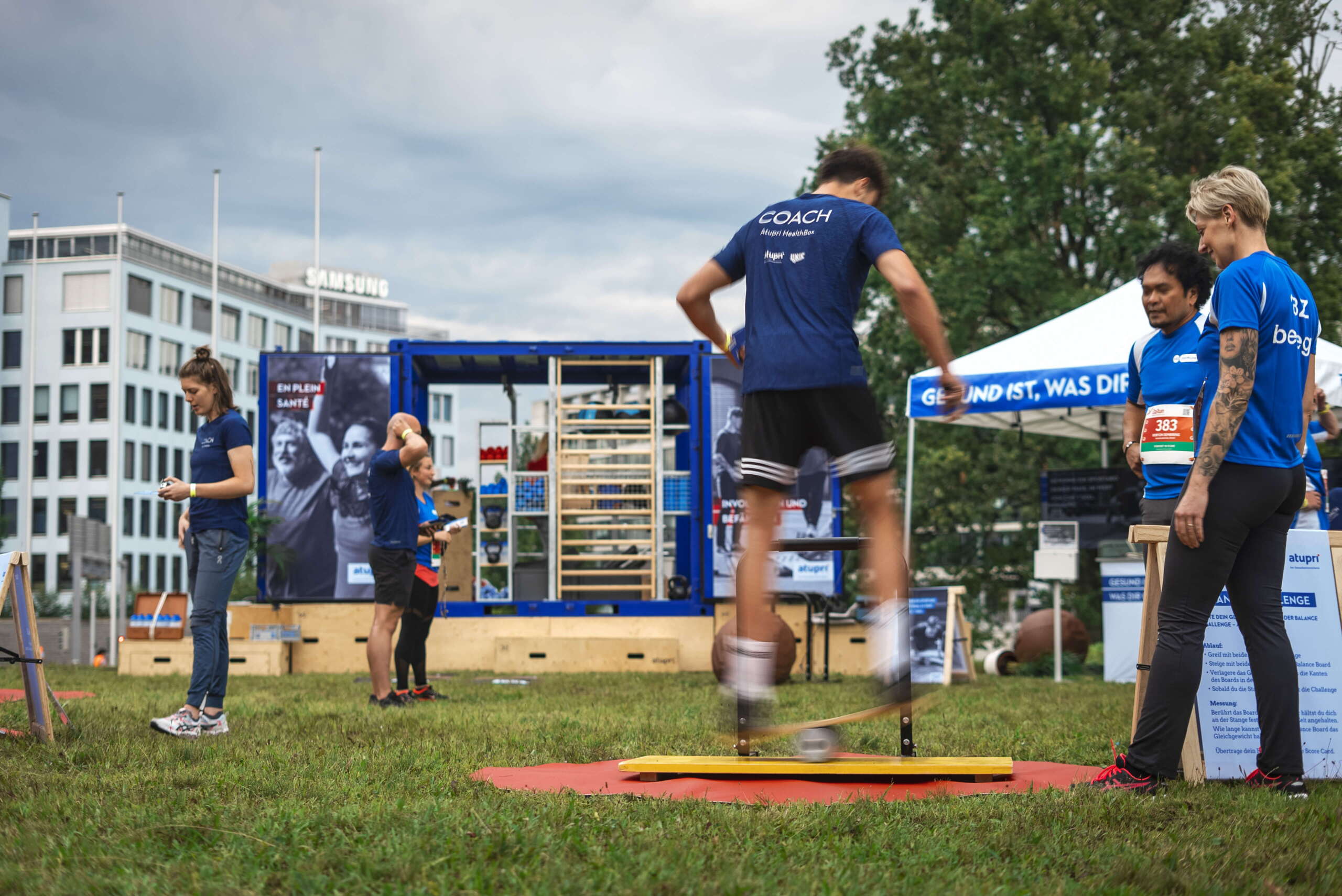 evenjo-ag-eventagentur-bern-atupri-healthbox.jpg Coach der Atupri Healthbox beim Koordinationstraining auf Balance-Board während Outdoor-Gesundheitsevent. Atupri Healthbox, Slackline, Koordinationstraining, Outdoor-Workout, Bewegung, Fitnessparcours, live kommunikation, Corporate-Event, Eventagentur bern, eventplanung, firmenevents, teamevents bern, jubiläumsfeier, networking anlass, mitarbeiteranlass, Technik, Public Events, Sport und Entertainment Events, markenerlebnis, Brand Experience