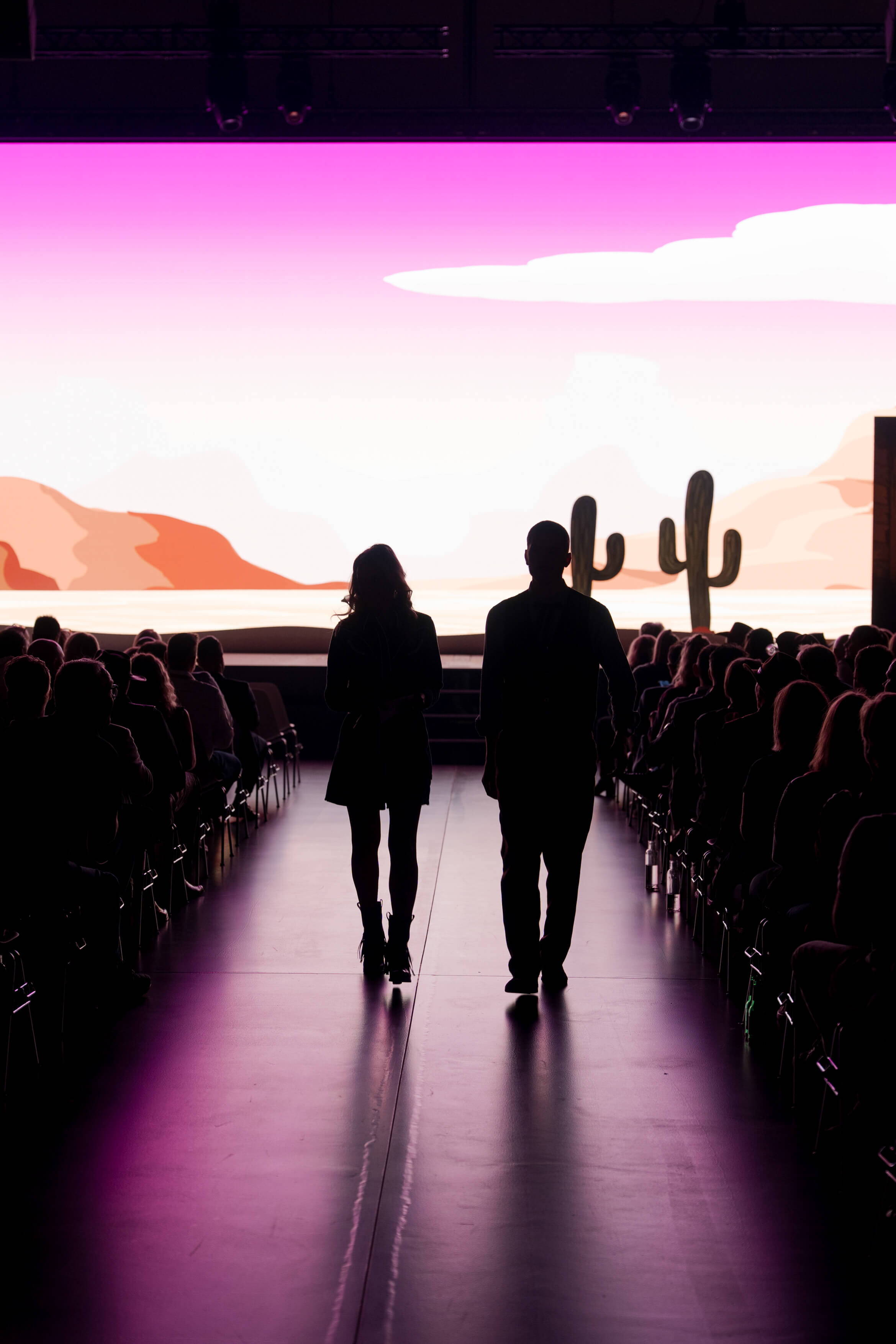 evenjo_xaveraward_preisverleihung_1.jpg Zwei Personen gehen durch das Publikum Richtung Bühne, im Hintergrund leuchtet ein pinker Western-Sonnenuntergang.
