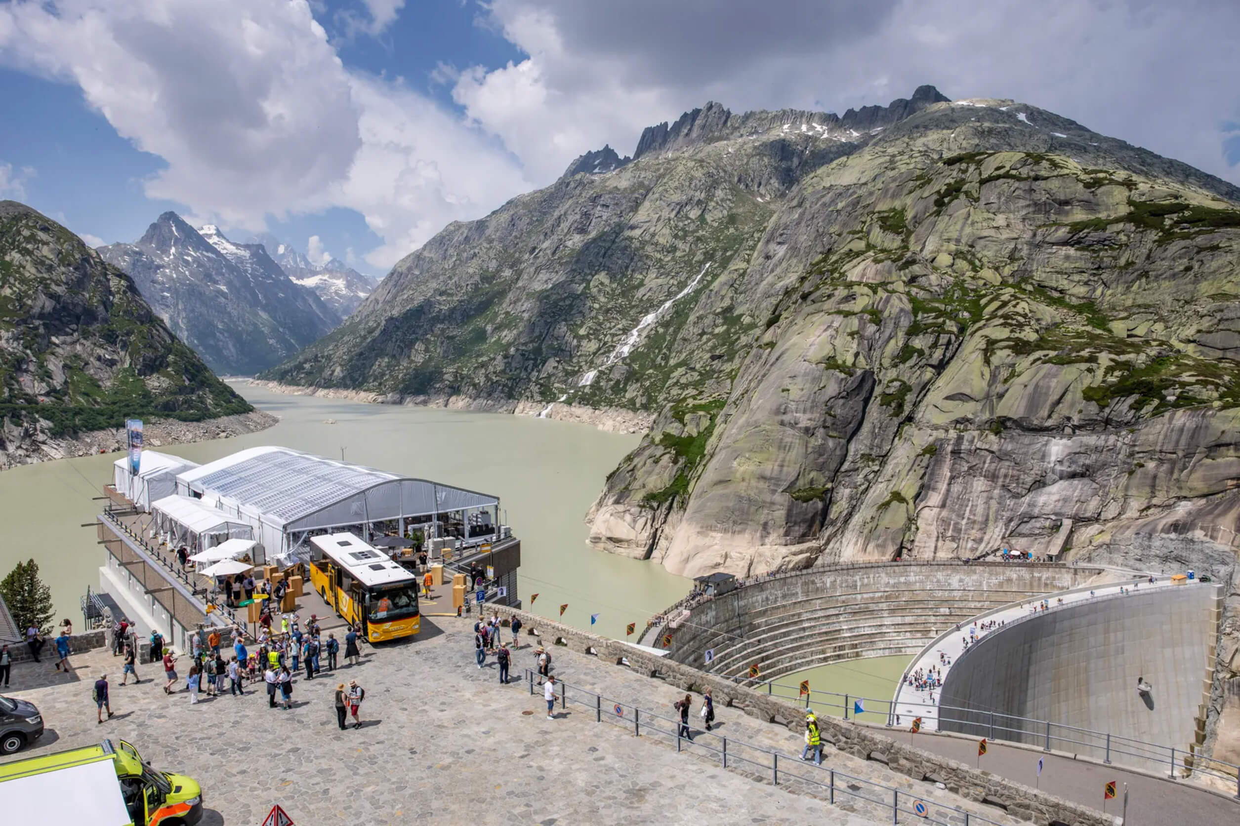 Jubiläumsveranstaltung der KWO auf der Grimsel-Staumauer: Gäste, Festzelt und Shuttlebus vor alpiner Bergkulisse und Stausee.