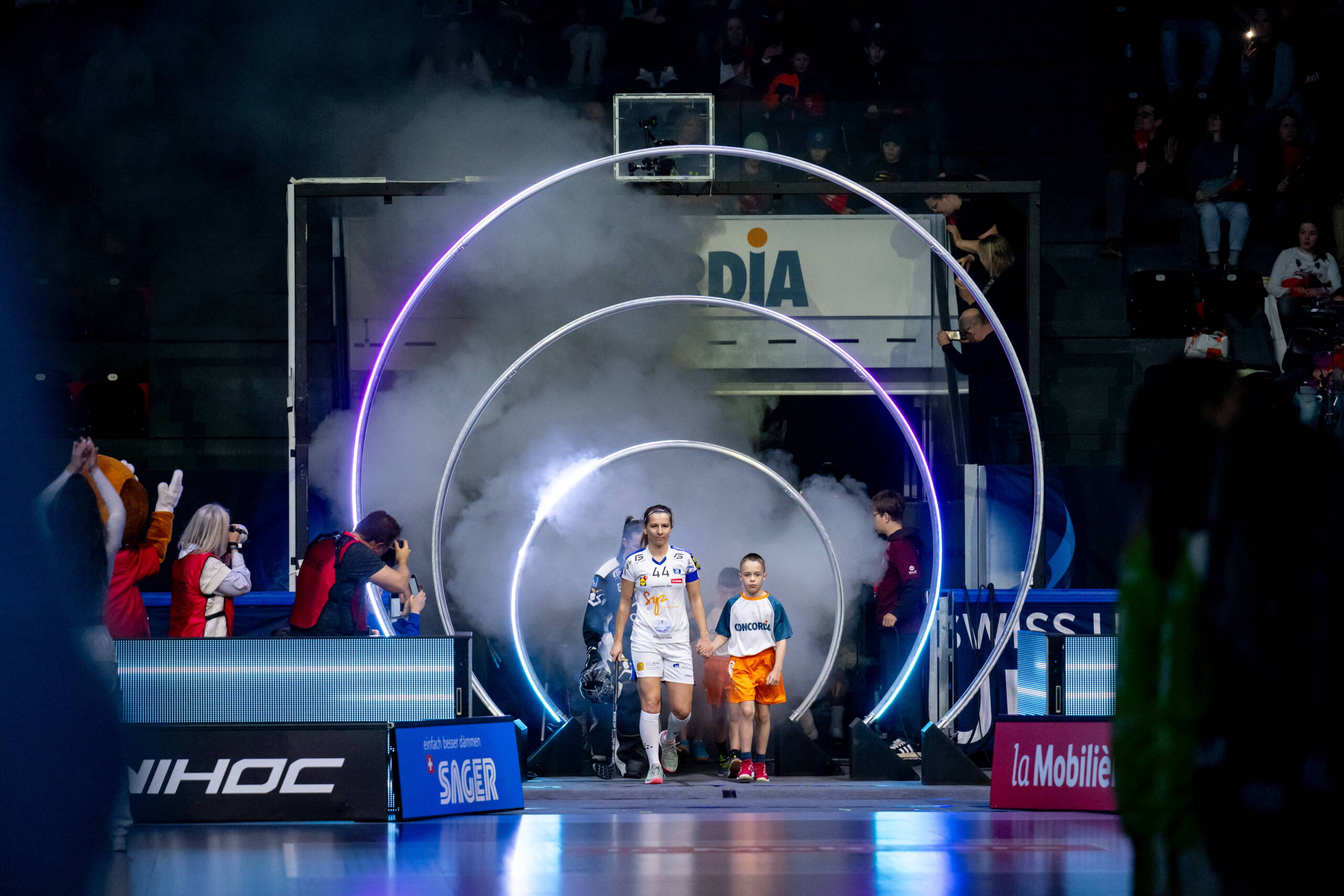 evenjo_sport-event_swiss-unihockey_superfinale_zug-united-unihockey-tigers-langnau.jpg Spielerinnen des Unihockey Superfinals betreten mit Kindern und unter Lichteffekten das Spielfeld, beeindruckender Einlaufmoment. Einlaufshow, Lichteffekte, Spielerinnen, Emotionen, Sportevent Schweiz, Floorball, live kommunikation, Corporate-Event, Eventagentur bern, eventplanung, firmenevents, teamevents bern, jubiläumsfeier, networking anlass, mitarbeiteranlass, evenjo-Team, Technik, Public Events, Sport und Entertainment Events, markenerlebnis, Brand Experience, Corporate-Event, Lichtshow, Eröffnungsshow