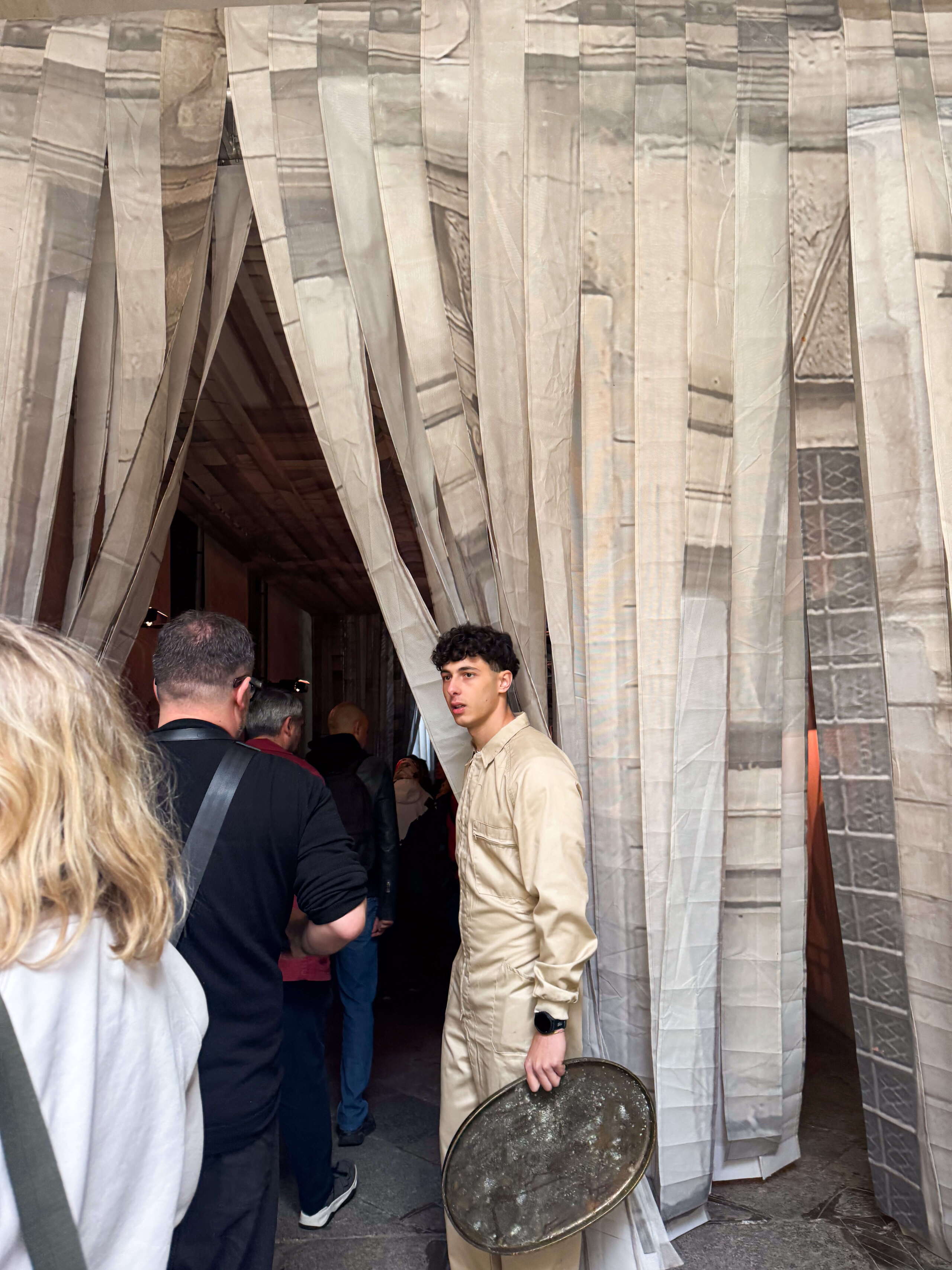 Besucher:innen betreten die immersive Aesop-Installation am Fuorisalone Milano durch halbtransparente Stoffbahnen. Eine Person in beiger Arbeitskleidung hält ein grosses Metaltablett und begleitet die Gäste in die Installation.