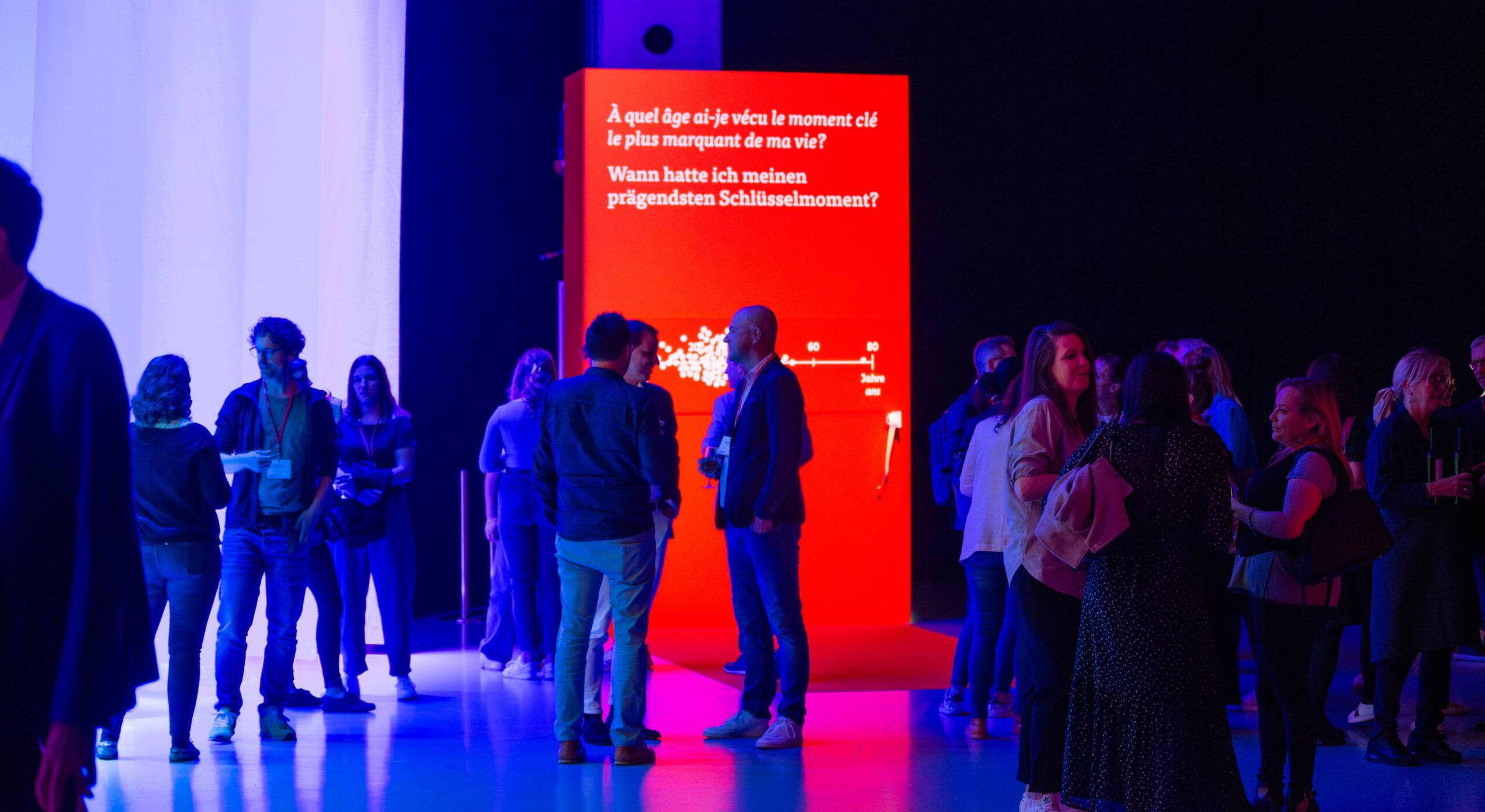 evenjo_mobiliar_avanti_corporateevent.jpg Besucher interagieren bei einem vor einer auffälligen roten Erlebnisinstallation mit der Frage nach prägenden Schlüsselmomenten. Eventtechnik, live kommunikation, markenerlebnis, Brand Experience, interaktive Ausstellung, Eventbeleuchtung, Corporate-Event
