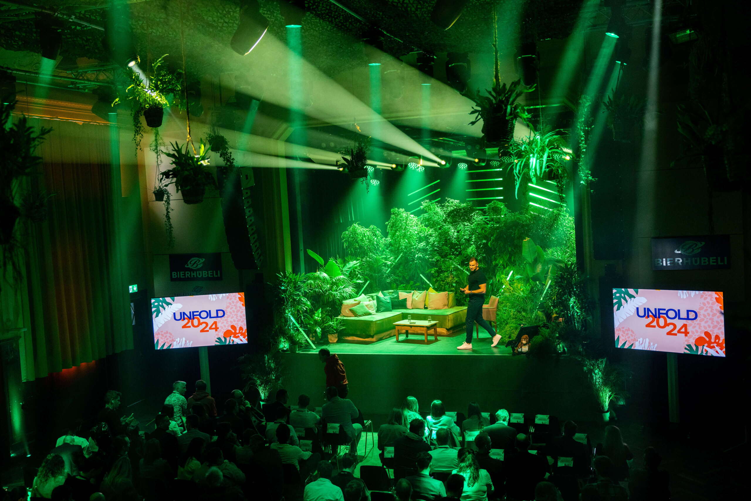 unfold_businessevent_evenjo_ag_bierhuebeli_bern-5.jpg Bühnenbild im Dschungel-Look mit grünem Licht und üppiger Bepflanzung beim Corporate Event Unfold 2024 von Cleverclip im Bierhübeli Bern. Corporate Event, Eventbühne, Eventdesign, grüne Lichtstimmung, Dschungel Deko, Business Event, live kommunikation, Eventagentur bern, eventplanung, firmenevents, teamevents bern, jubiläumsfeier, networking anlass, mitarbeiter:innenanlass, Technik, Public Events, Sport und Entertainment Events, markenerlebnis, Brand Experience