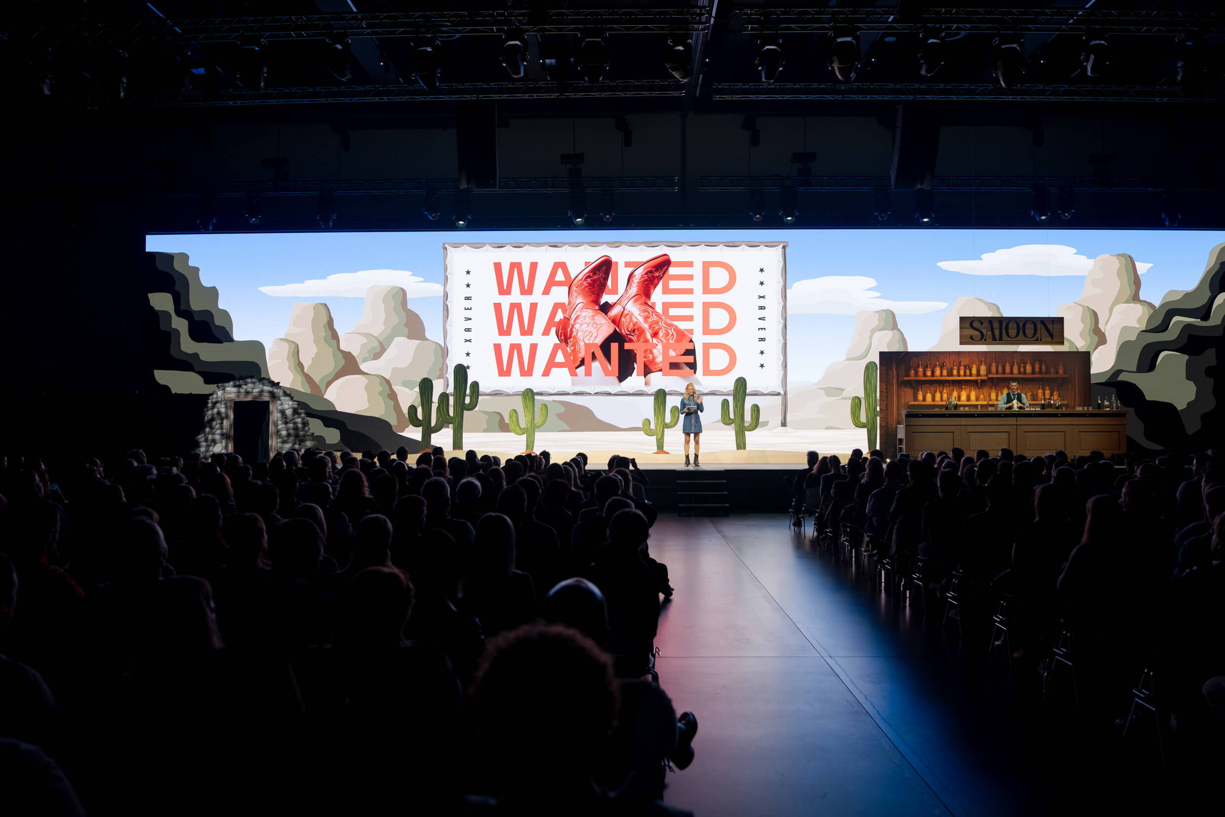 evenjo_xaveraward_preisverleihung.jpg Blick auf die Bühne der XAVER Award Show 2025 im Western-Stil. Auf der Leinwand steht «WANTED», die Moderatorin begrüsst das Publikum.