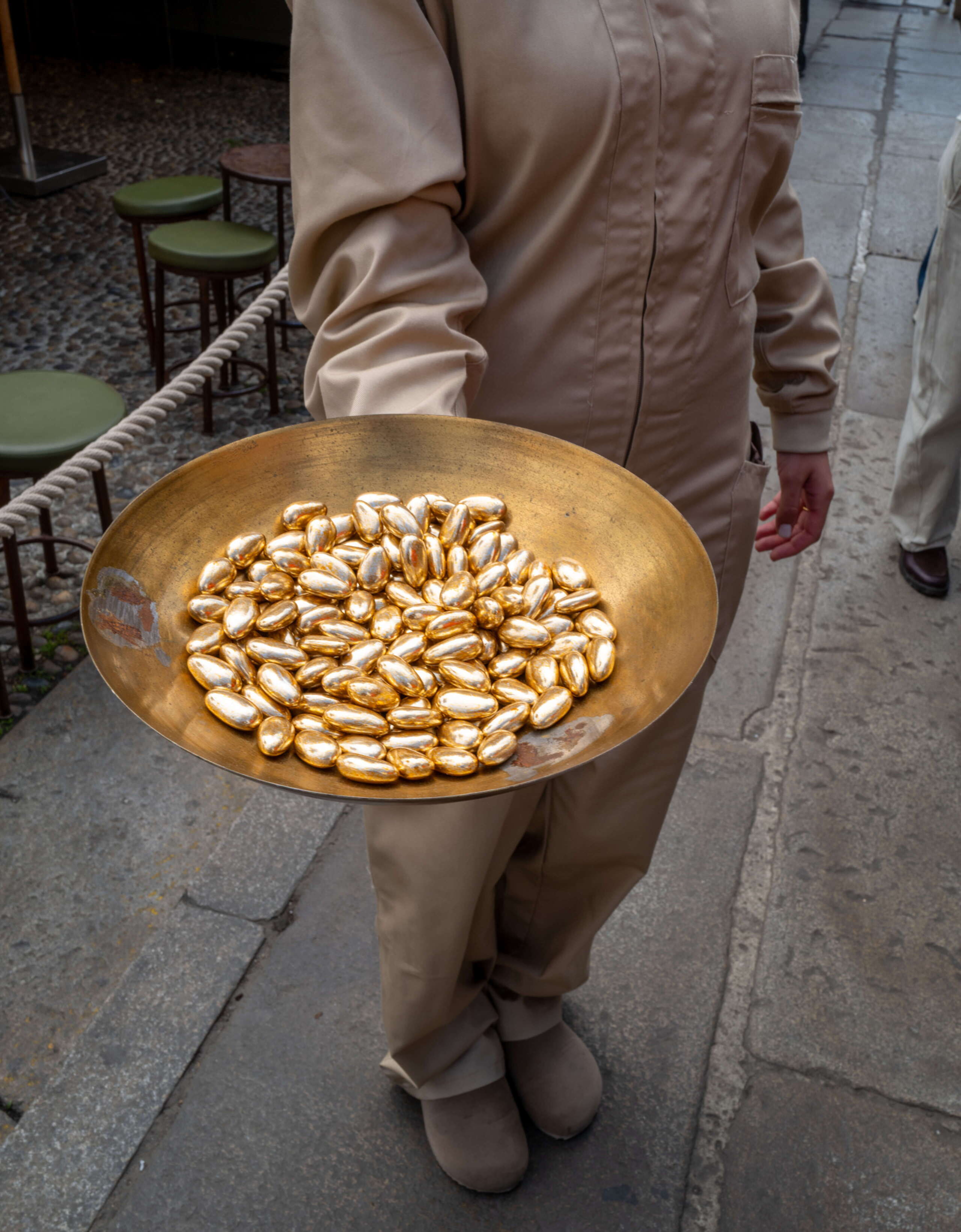 Eine Person in beiger Arbeitskleidung trägt bei der Aesop-Installation am Fuorisalone der Milan Design Week in Milano eine grosse goldene Schale mit glänzenden, mandelförmigen Objekten. Die Szene wirkt wie Teil einer performativen Begrüssung.